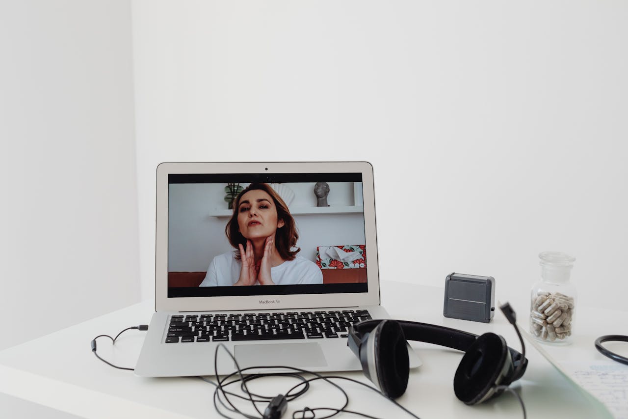 A woman conducting a telemedicine session visible on a laptop screen with headphones nearby.